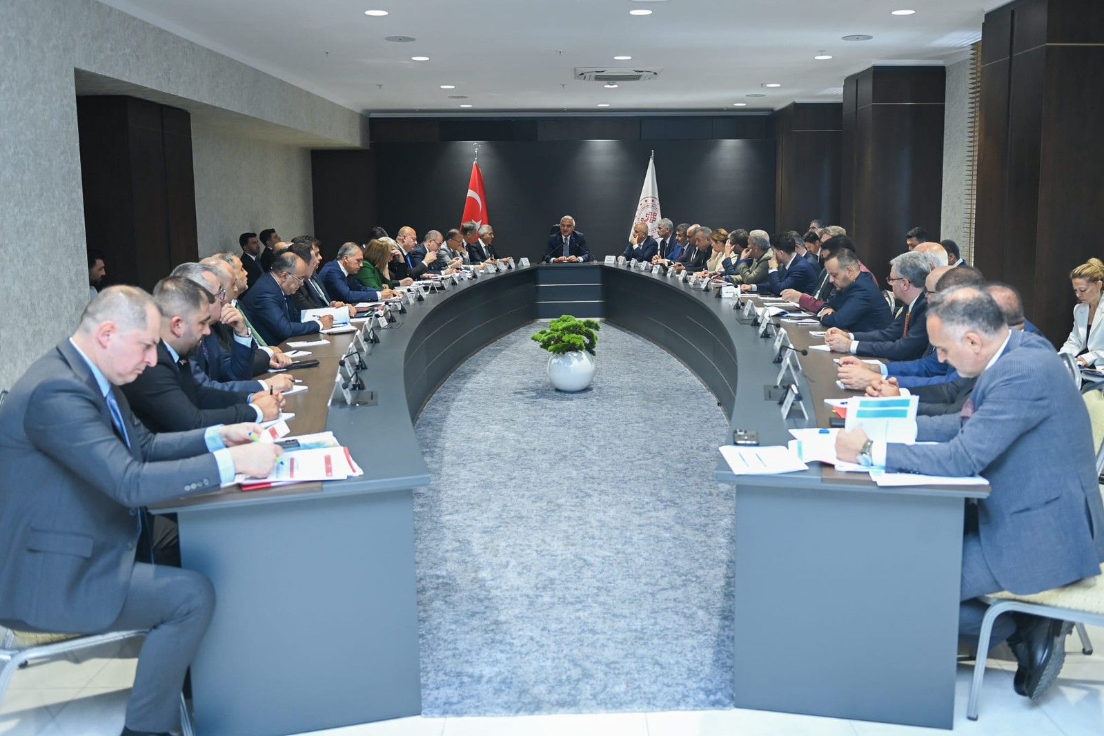U biçimli konferans masasında iki sıra resmî katılımcı oturuyor.