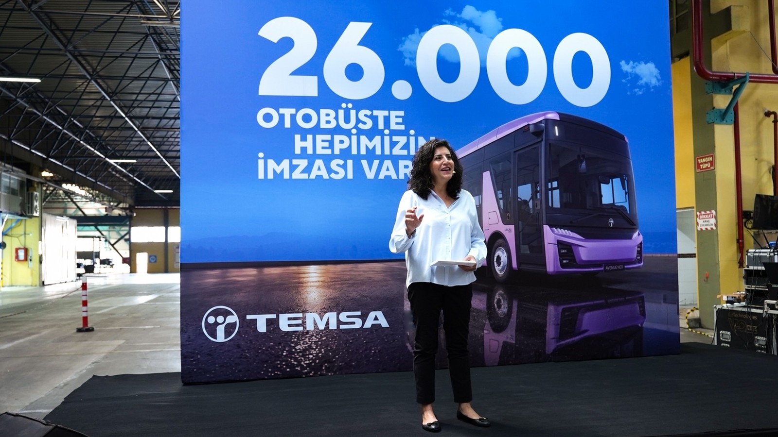 Beyaz gömlekli bir konuşmacı, '26.000 otobüste hepimizin imzası var' yazılı dev perde ve mor Avenue Neo görseli önünde sahnede