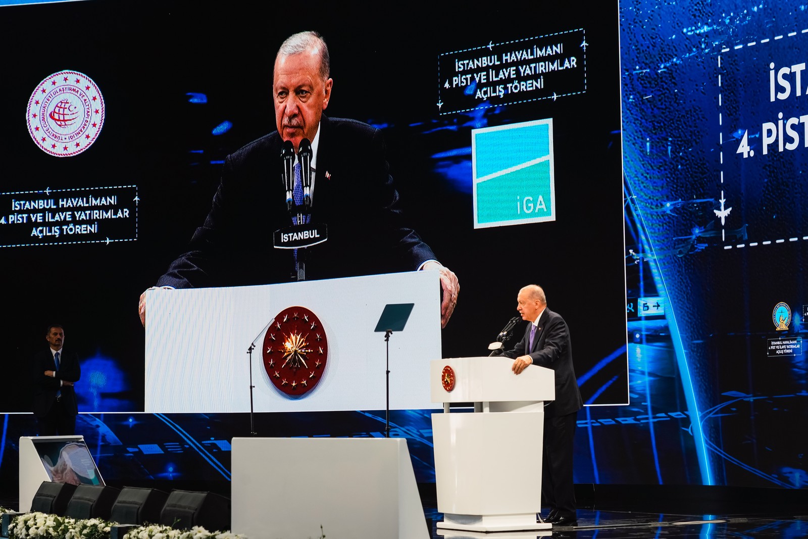 Havalimanı pist açılışında kürsüde konuşan resmî temsilci ve sahne ekranı.
